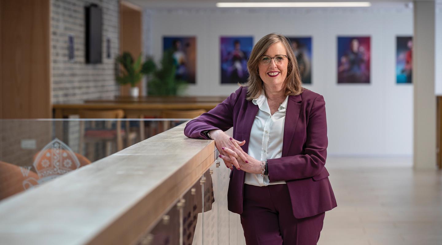 Melanie is smiling, dressed in a smart pink suit, and stood in a reception area with art on the wall behind her.