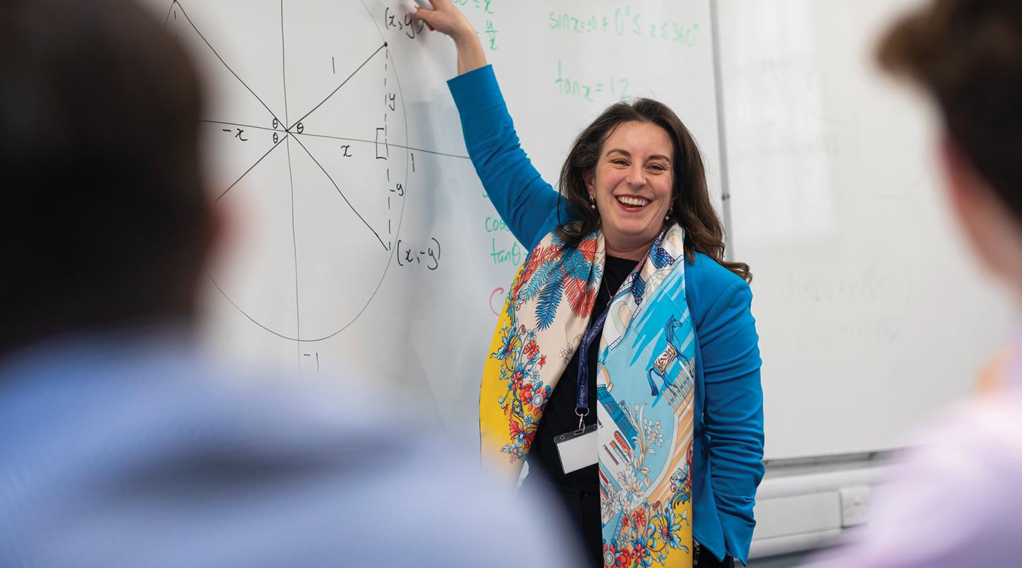 The headmaster, dressed in a smart blue jacket, is smiling and pointing at the whiteboard, with students in the foreground.