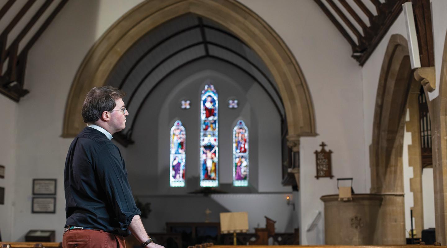 The reverend is stood inside his church looking thoughtful, with stained glass windows in the background. 