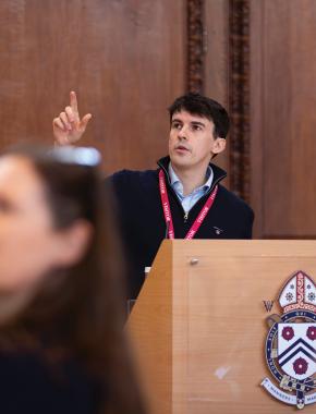 A man with dark hair and blue shirt, giving a speech at Winchester College.