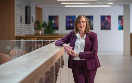 Melanie is smiling, dressed in a smart pink suit, and stood in a reception area with art on the wall behind her.