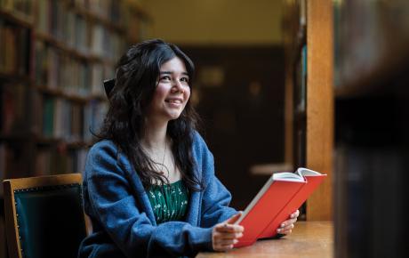 Michelle, with dark hair and wearing a green dress with blue cardiagn, is smiling whilst holding a book in the library