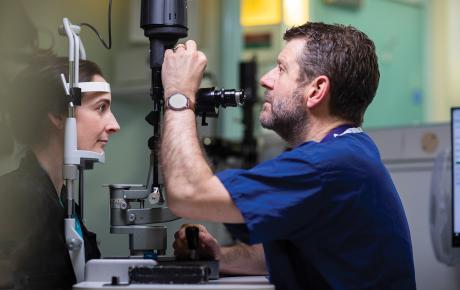 Gus, wearing blue hospital uniform, is doing an eye test on a patient.