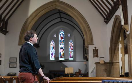 The reverend is stood inside his church looking thoughtful, with stained glass windows in the background. 