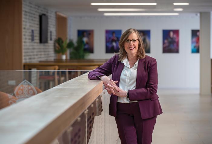 Melanie is smiling, dressed in a smart pink suit, and stood in a reception area with art on the wall behind her.