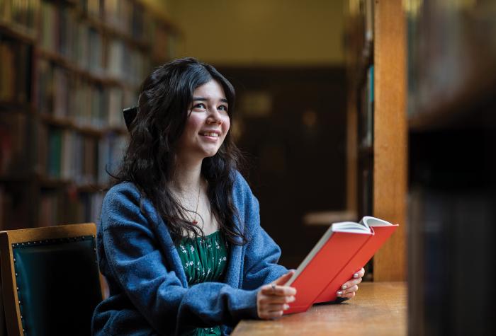 Michelle, with dark hair and wearing a green dress with blue cardiagn, is smiling whilst holding a book in the library