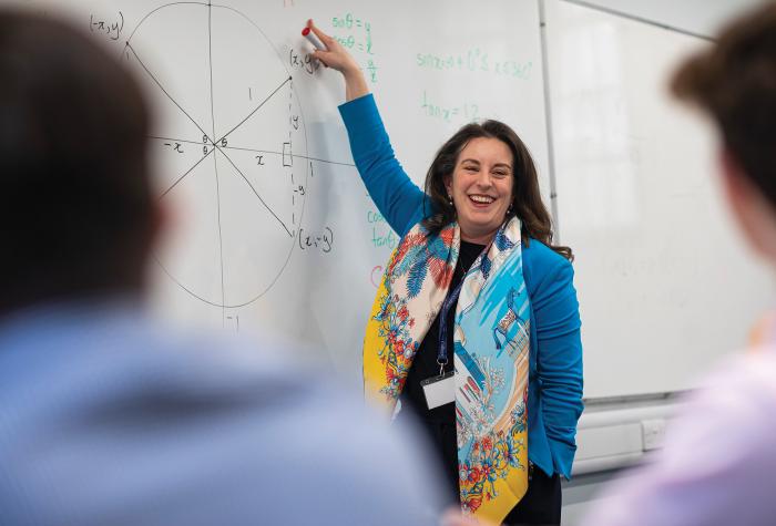 The headmaster, dressed in a smart blue jacket, is smiling and pointing at the whiteboard, with students in the foreground.