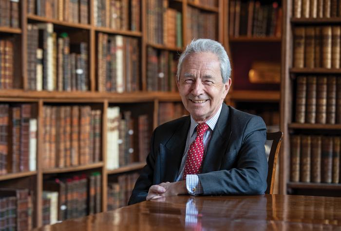 A smartly dressed man is smiling at the camera, sat at a desk with bookshelves in the background.