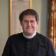 The Reverend - with glasses, dark hair and wearing a black shirt - is smiling at the camera.